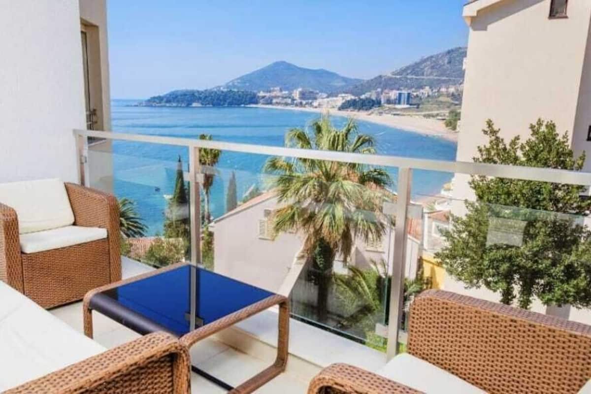 Uma varanda com mobiliário de vime, uma mesa de vidro e vista para o mar e montanhas.