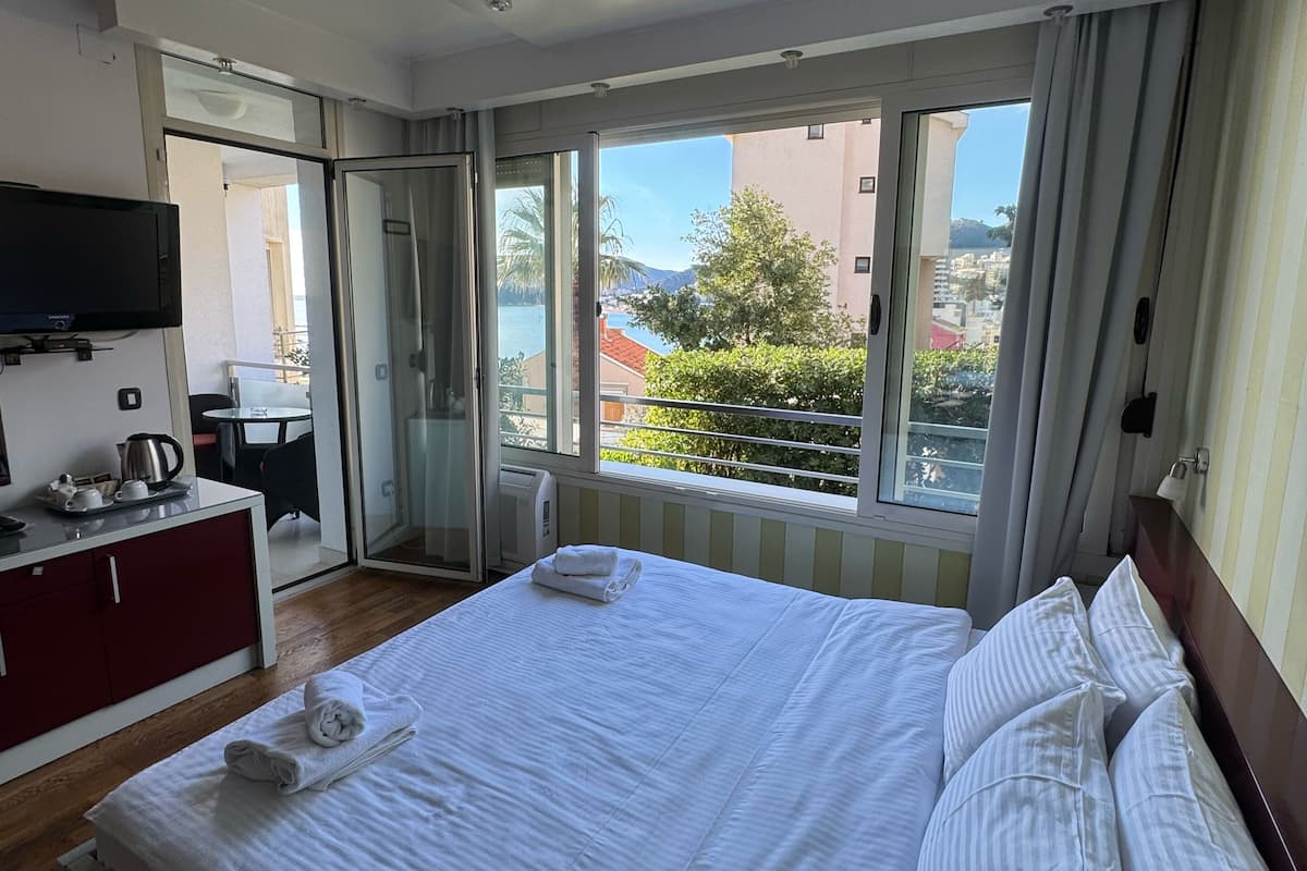 Quarto de hotel com uma cama grande, televisão presa à parede, escrivaninha com chaleira e vista da cidade através de grandes janelas.