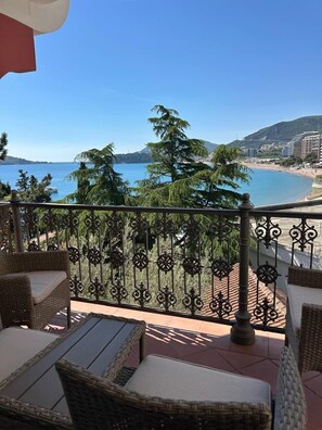 Ein Balkon mit Korbmöbeln, einem Metallgeländer und Blick auf Strand und Berge.