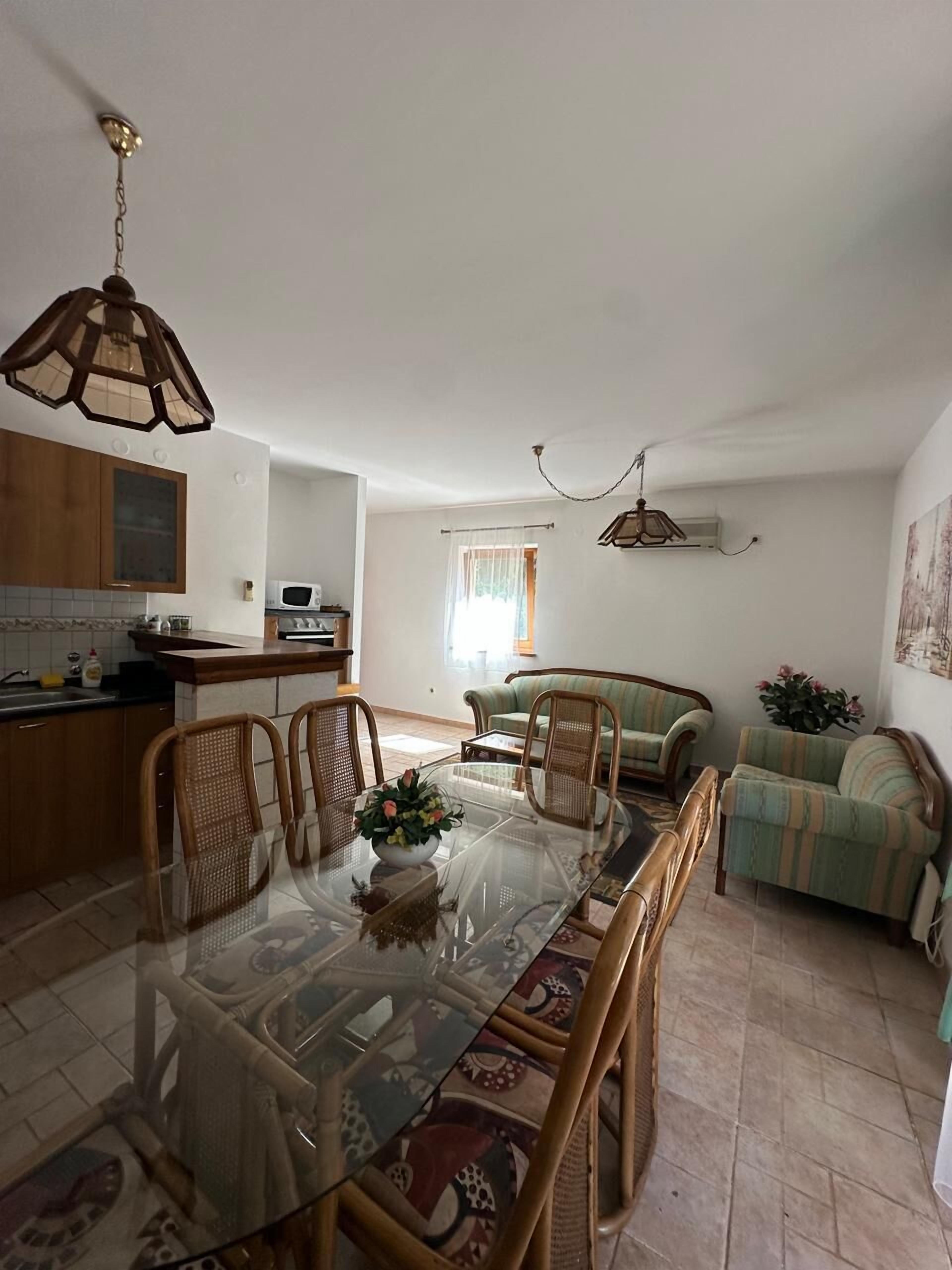 Ein Essbereich mit Glastisch und Korbstühlen, eine Küche mit Holzausstattung und ein Wohnzimmer mit Sofa und einem gemusterten Teppich.