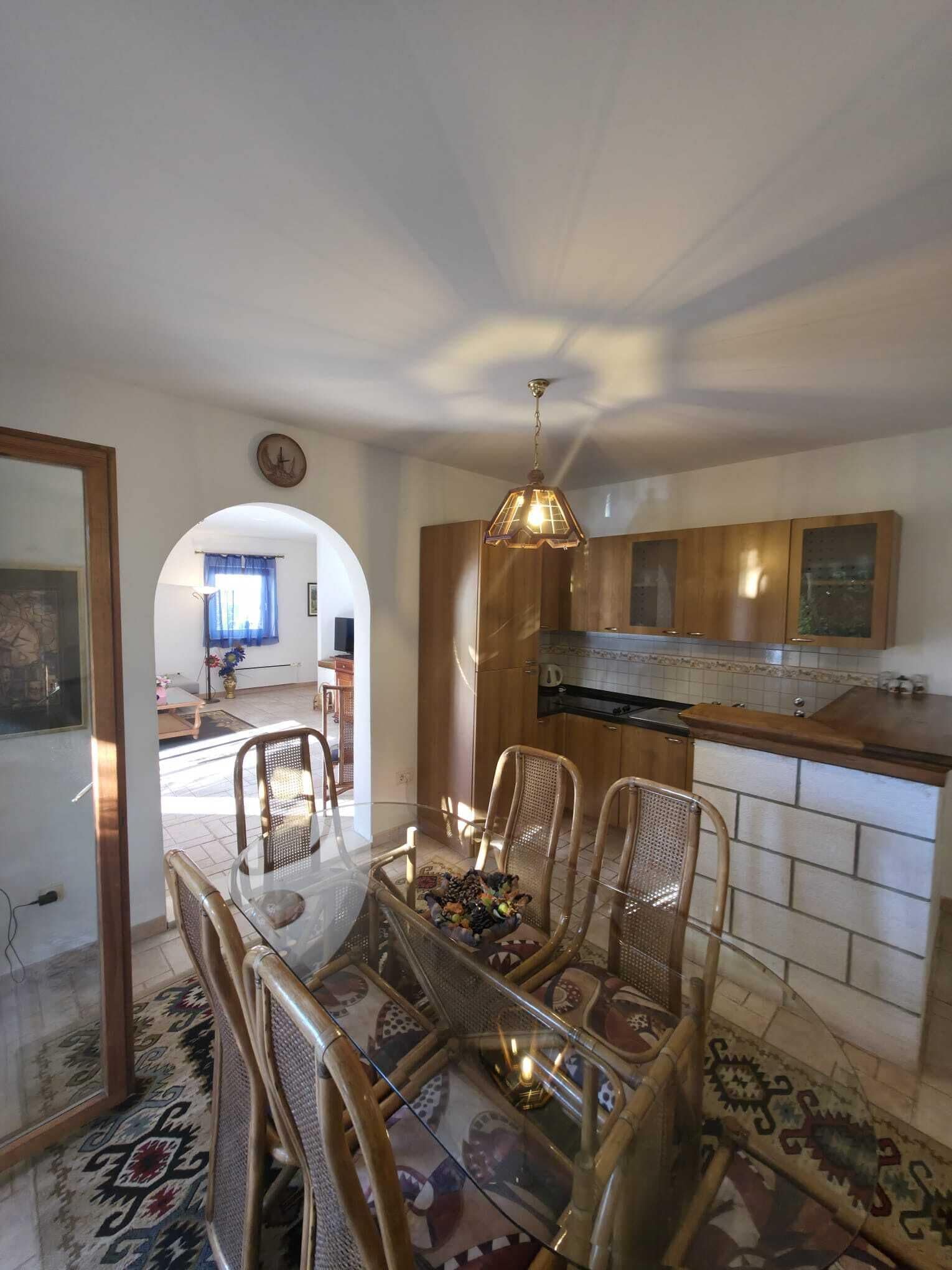 A dining area with a glass table and wicker chairs, a kitchen with wooden cabinets, and a visible clock on the wall.