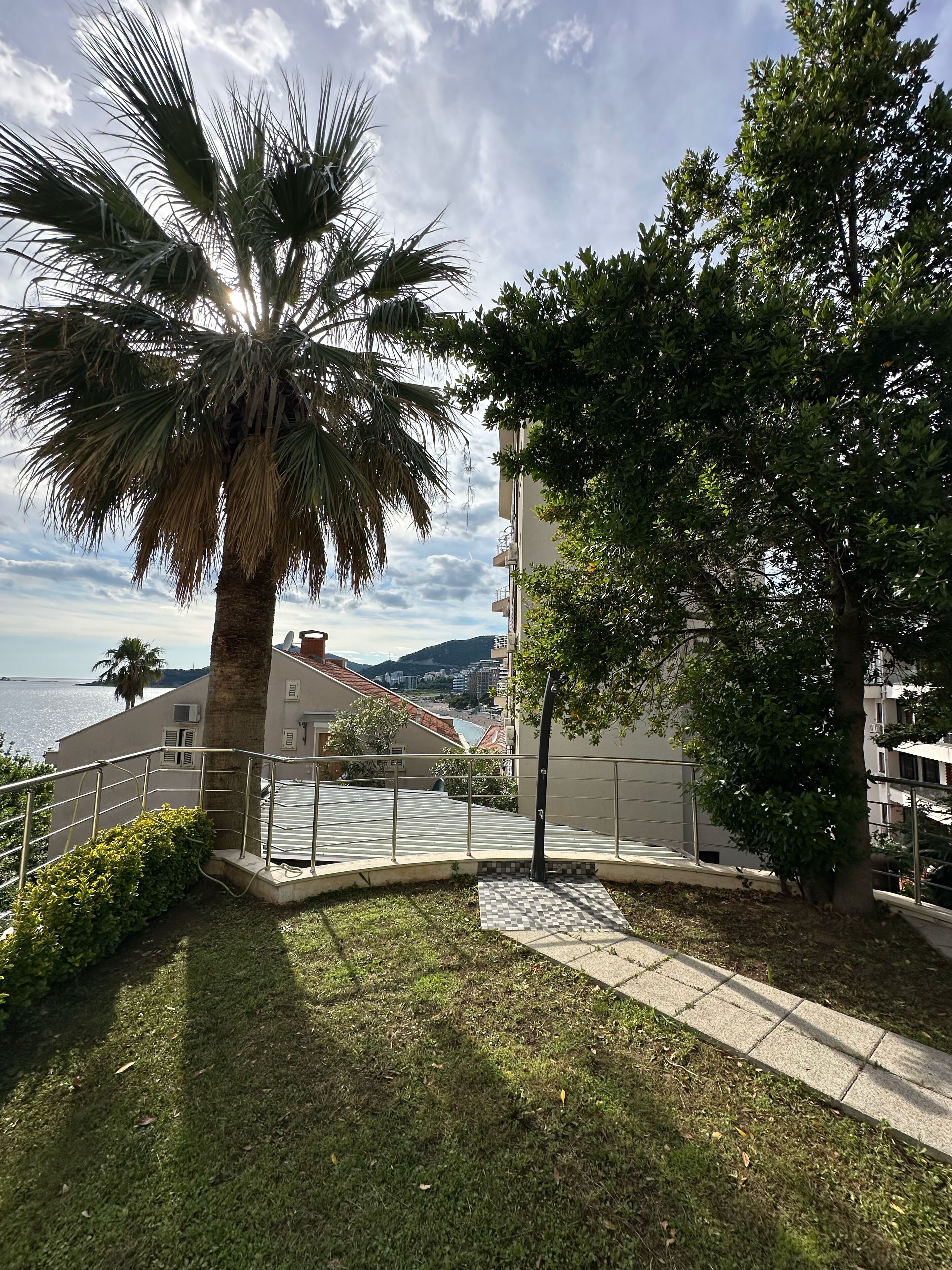 A balcony with a view of the sea, palm trees, and a tiled pathway.