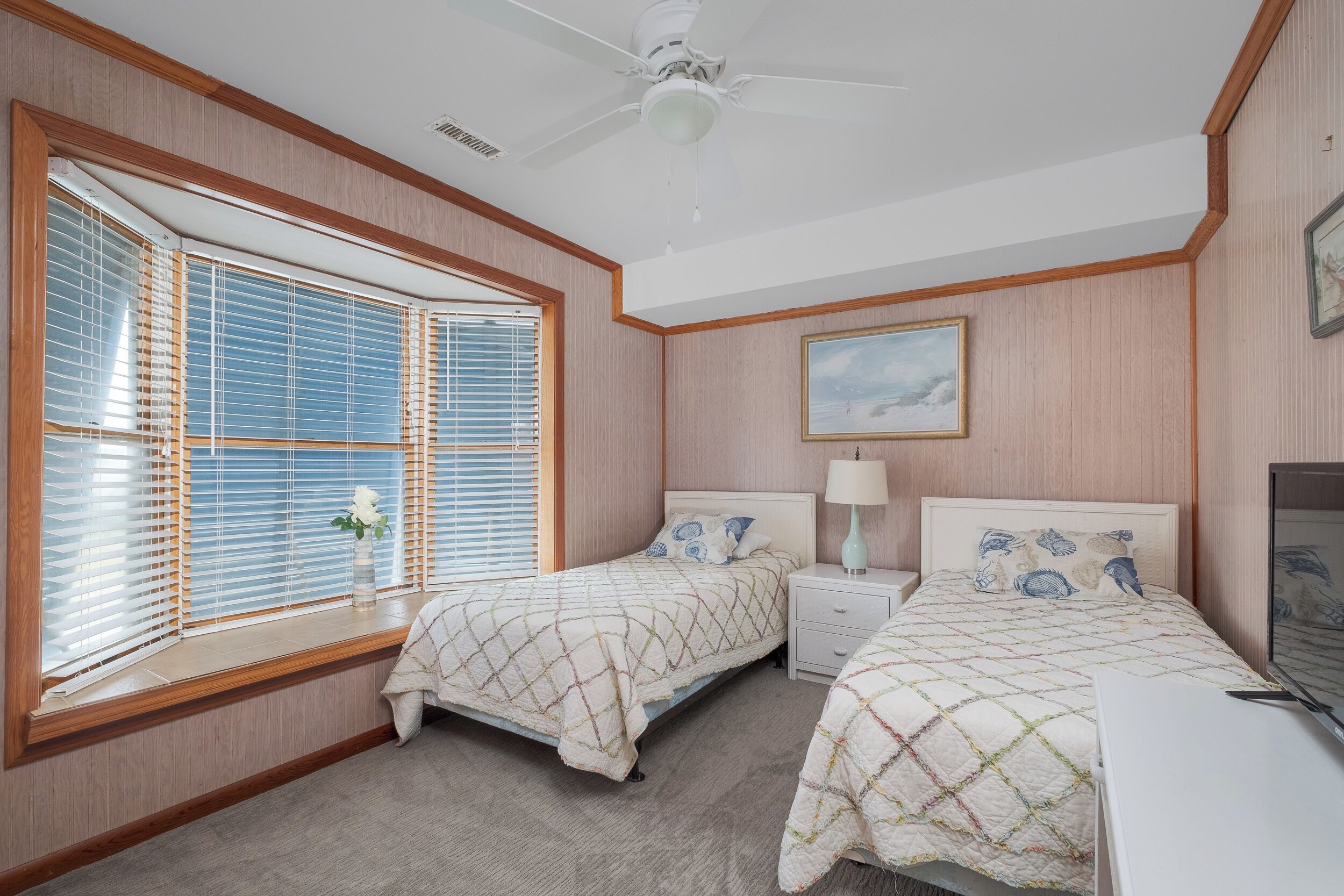 A bedroom with two beds, a ceiling fan, a nightstand with a lamp, a TV, and a window with blinds.