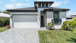 A modern two-story house with a garage, a paved driveway, and landscaped garden with small plants and a palm tree. - 1 Mile to Beach! | Heated Pool | New Construction (Naples)