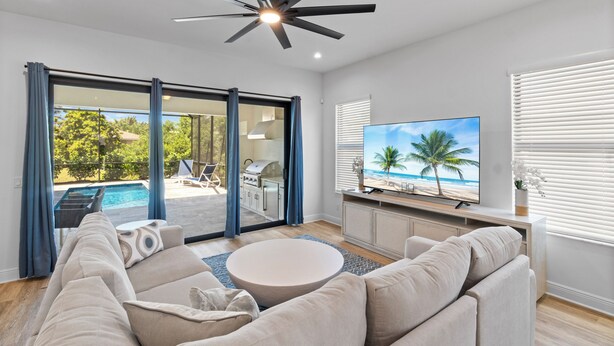 A modern living room with a large TV, comfortable seating, and a view of a pool and outdoor area. - 1 Mile to Beach! | Heated Pool | New Construction (Naples)