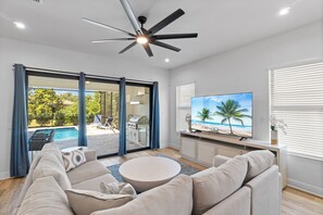 A modern living room with a large TV, comfortable seating, and a view of a pool and outdoor area. - 1 Mile to Beach! | Heated Pool | New Construction (Naples)