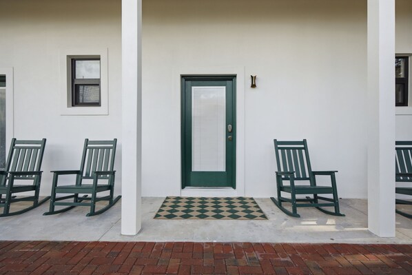 A building entrance with a green door, two rocking chairs, a small window, and a doormat.