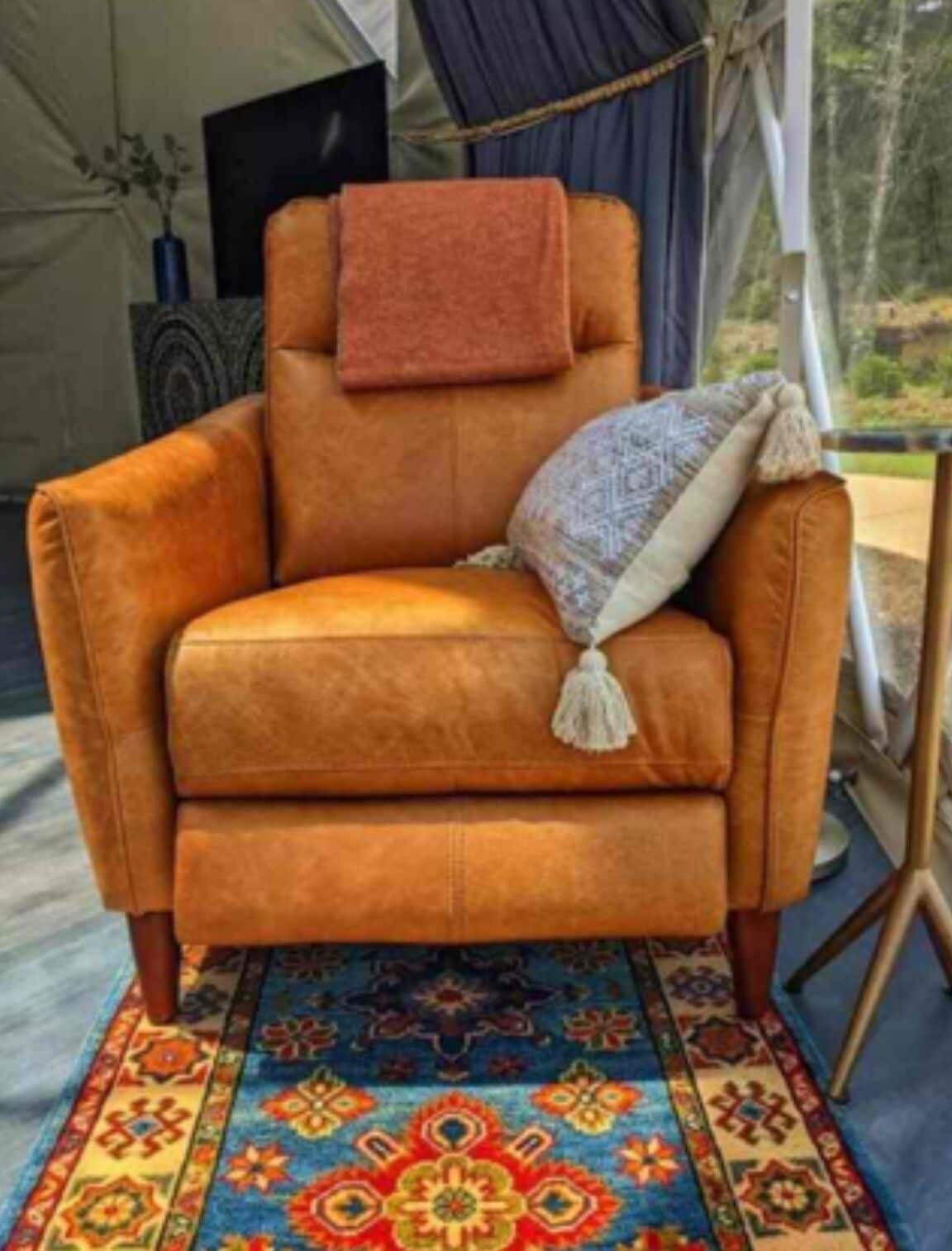Ein gemütlicher orangefarbener Sessel mit einer Decke und einem Zierkissen, platziert auf einem gemusterten Teppich in einer Zelteinrichtung im Freien.
