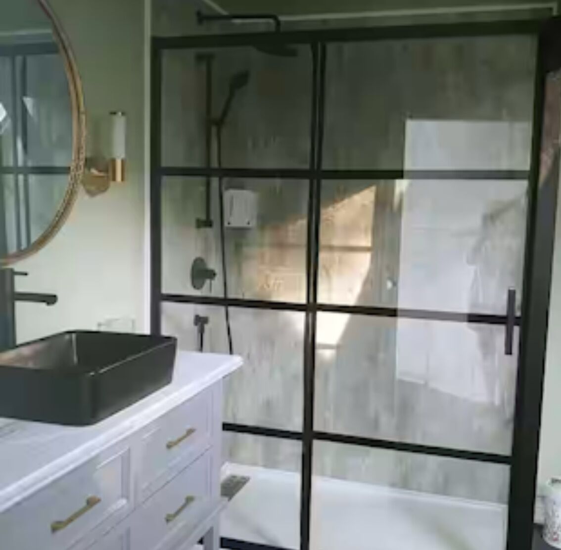 Ein Badezimmer mit Duschkabine aus Glas, einem weißen Waschtisch mit schwarzem Becken und einem Spiegel mit goldener Rahmung.