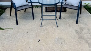 Un patio con dos sillas, una mesa, una puerta blanca y una ventana con persianas.