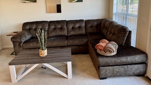 Una sala de estar con un sofá seccional marrón oscuro, una mesa de centro de madera con una cesta tejida y una planta, y dos cuadros abstractos colgados en la pared.