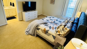 Un dormitorio con cama, televisor, ventilador de techo, ventana con cortinas y un baño visible a través de una puerta abierta.