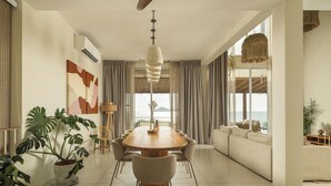 Un espace de repas moderne avec une table et des chaises en bois, une plante en pot et une vue sur l’océan grâce à de grandes fenêtres.