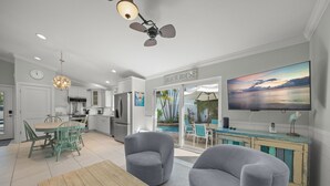 A modern kitchen with white cabinets, a dining area with teal chairs, and a large TV on the wall.