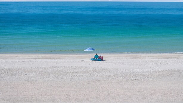 A beach with a clear blue sea, a sandy shore, and a person sitting under a striped umbrella.