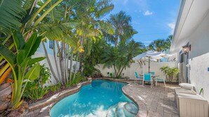 A swimming pool surrounded by tropical plants, a patio with chairs, and a white building.