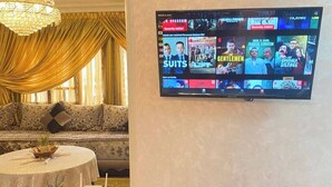 A living room with a flat-screen TV mounted on the wall displaying various streaming services. There is a round table with a white tablecloth and a potted plant. A chandelier hangs from the ceiling, and a sofa with a patterned fabric is visible in the background. - Charming 2-bedroom apartment with WiFi, AC in superb Fès (Fès)