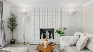 Una sala de estar con chimenea de ladrillo blanco, dos lámparas de pared, una mesa de centro de madera y un sofá con cojines.