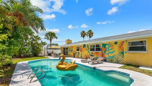 Una piscina con un flotador con forma de cisne, rodeada de palmeras y un mural colorido.