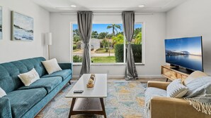 Una sala de estar con un sofá color turquesa, una mesa de centro, un televisor y vistas a una zona residencial con palmeras.