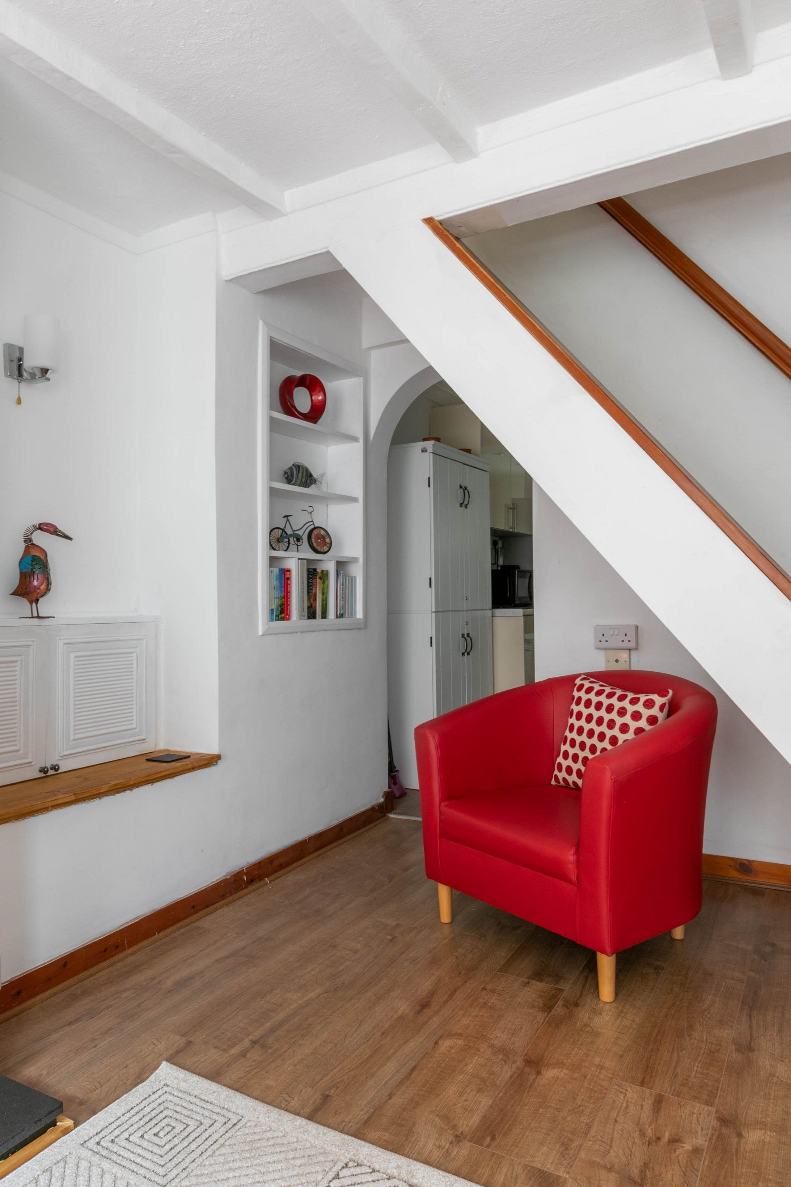 Una sala moderna con un sillón rojo, un estante blanco con objetos decorativos y una escalera que lleva a otra habitación.