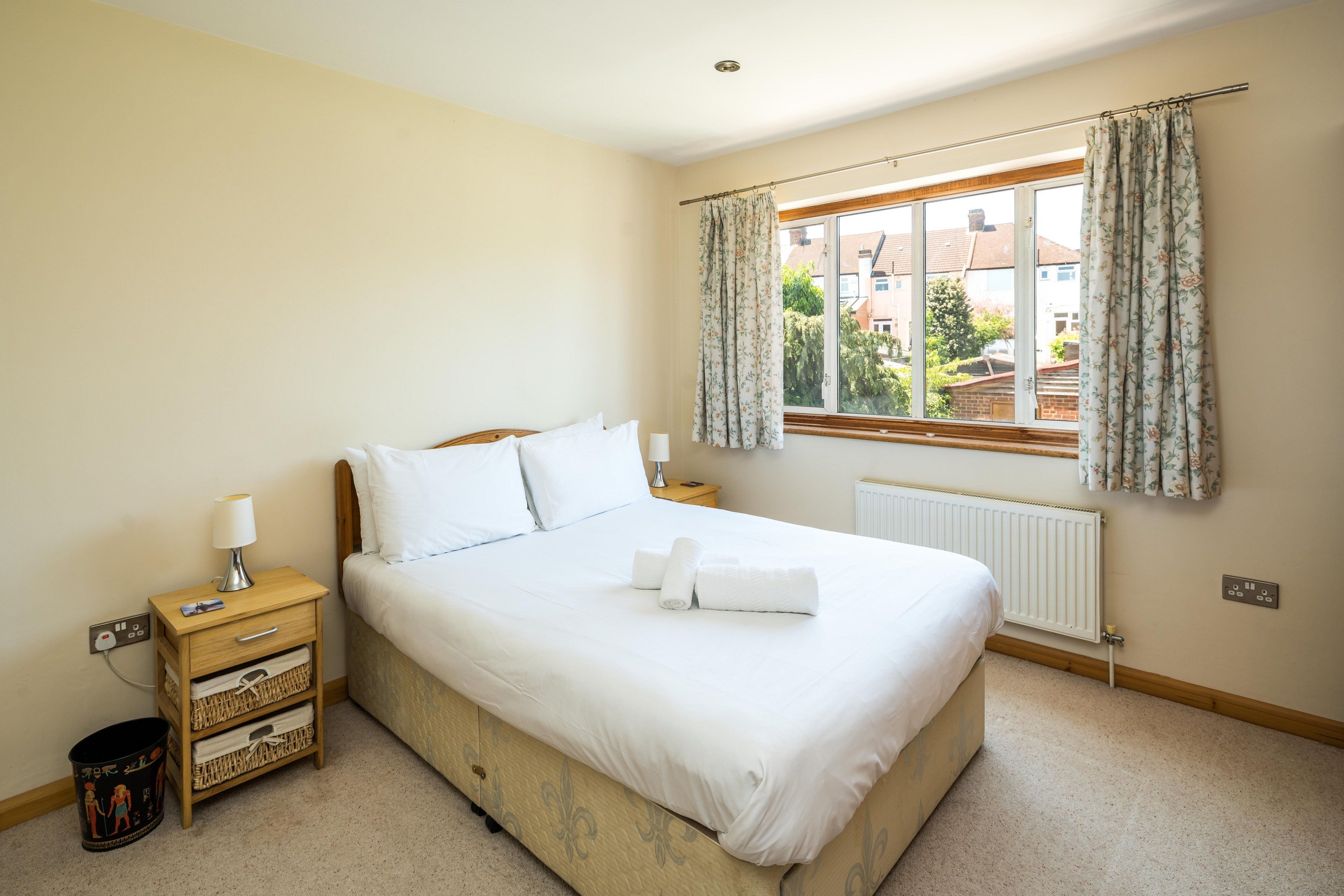 A neatly made bed with white linens, a bedside table with a lamp, a small trash bin, and a window with curtains.