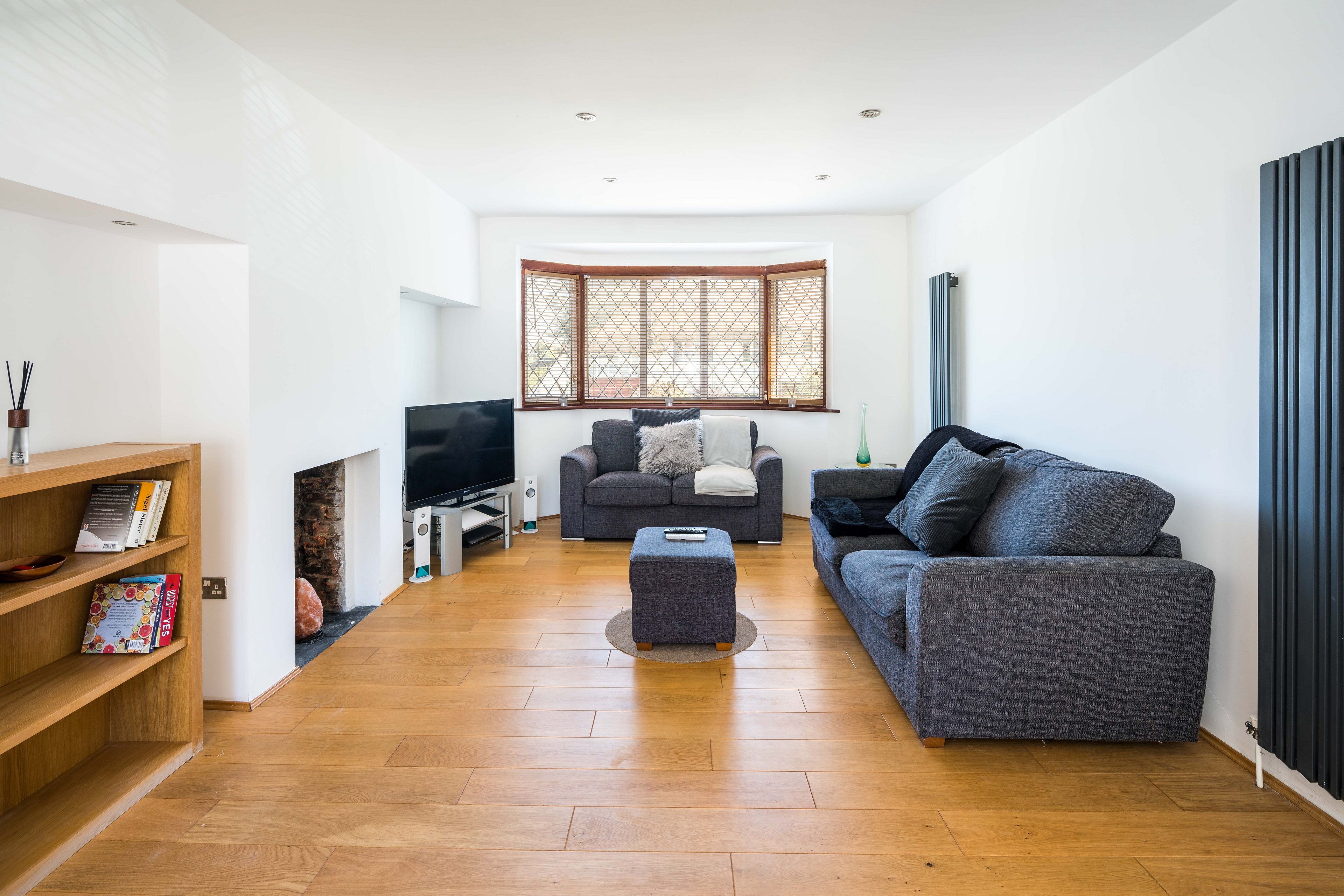 A modern living room with a wooden floor, two grey sofas, a small ottoman, a TV, and a bookshelf.