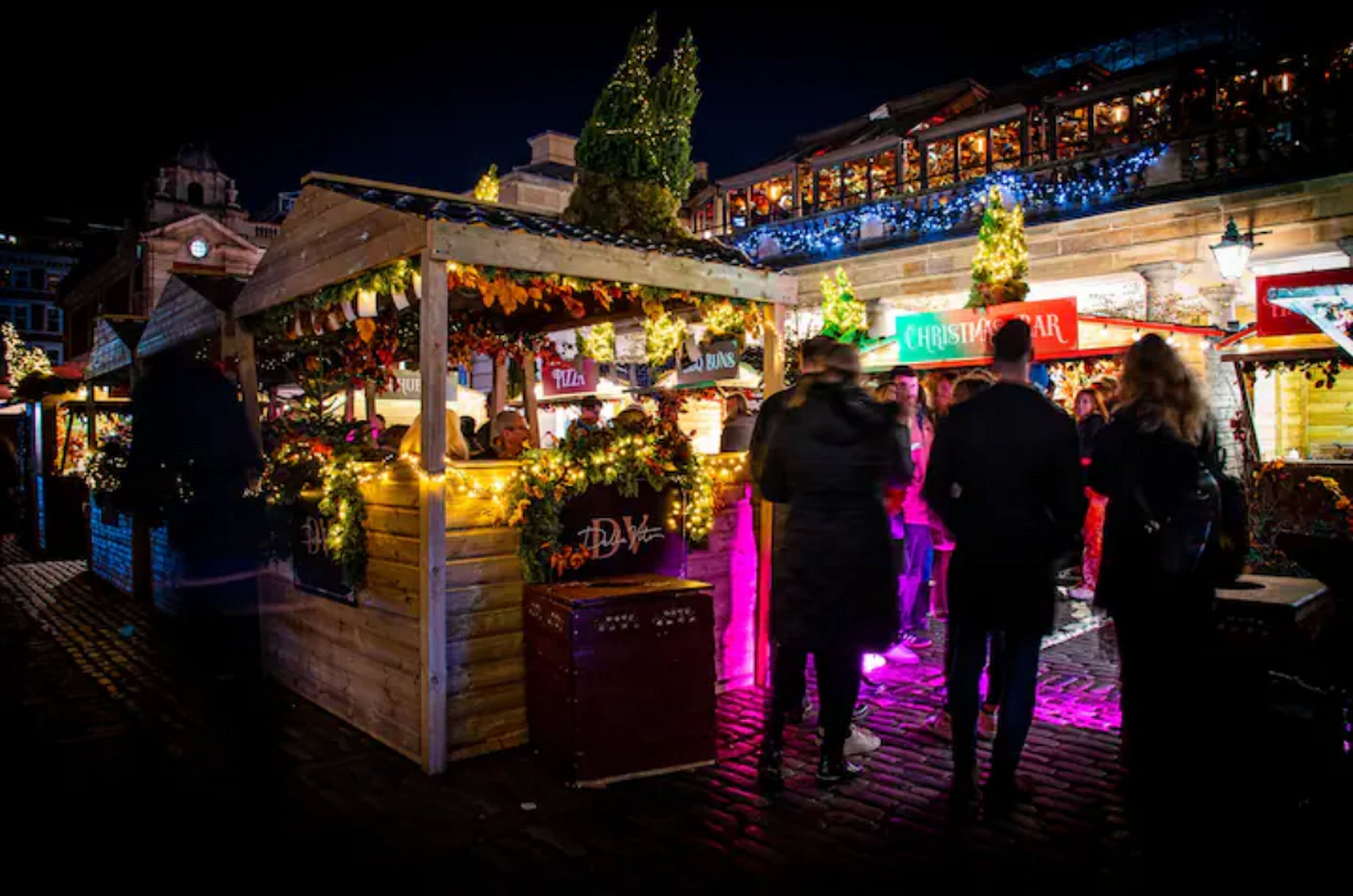 A festive outdoor market with wooden stalls, Christmas decorations, and a crowd of people.