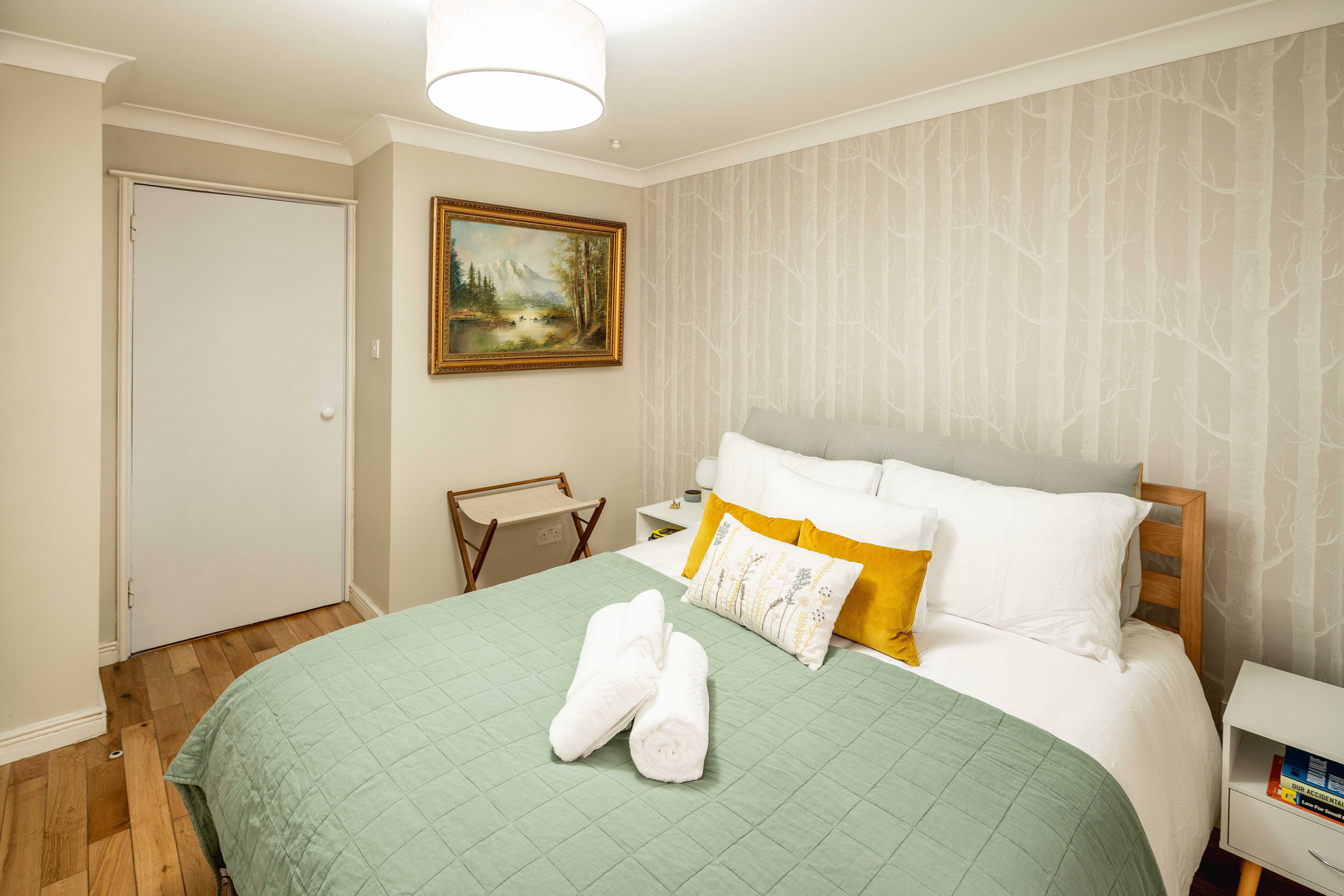 Una cama bien hecha con colcha verde, almohadas blancas y un cojín de acento amarillo. Hay un cuadro en la pared y una pequeña silla de madera.
