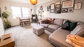 Ein Wohnzimmer mit einer Couch, einem Hocker und einem Esstisch mit Stühlen. An der Wand hängen gerahmte Bilder und in der Ecke steht eine Pflanze.