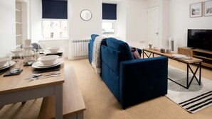 A modern living room with a blue sofa, a dining table set with white plates and glasses, a television, and a large window with blinds. - Historic 2Bd Maisonette In Coastal Eyemouth (Eyemouth)