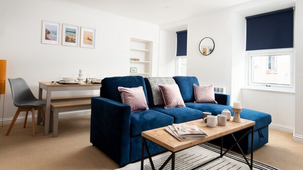 A modern living room with a blue sofa, a wooden coffee table, and a dining area with a table and chairs. - Historic 2Bd Maisonette In Coastal Eyemouth (Eyemouth)