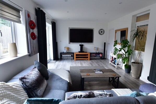 A modern living room with a grey sofa, a coffee table, and a TV. There is a bookshelf with books and a potted plant. - 2Bd Garden Home - A Short Walk To Stonehenge (Salisbury)