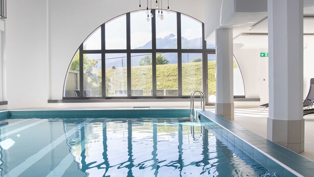 An indoor swimming pool with clear blue water, large windows offering a view of greenery and mountains, and a metal ladder for entering the pool. - La Siala Apt.51 by Interhome (Falera)