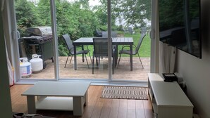 A room with a sliding glass door leading to an outdoor patio. Inside, there is a white bench, a small table, and a flat-screen TV mounted on the wall.
