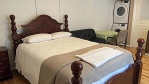 A neatly made bed with a wooden headboard, a bedside table, and a washing machine in the background. - Garden Annex (North Garden)