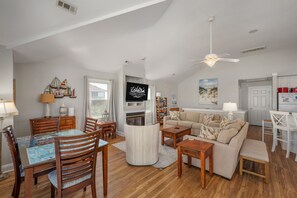 A spacious living room with a high ceiling, a dining area with wooden chairs and a glass table, and a kitchen area with a refrigerator and a white cabinet. - Gone Coastal: Private Pool, Hot Tub, Ocean and Sunset Views! (Nags Head)