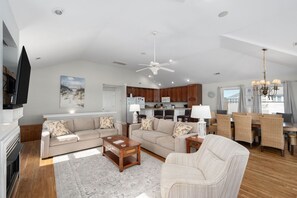 A spacious living room with a fireplace, two sofas, a coffee table, and a dining area with a chandelier. - Gone Coastal: Private Pool, Hot Tub, Ocean and Sunset Views! (Nags Head)
