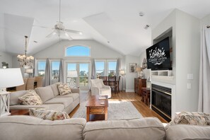 A spacious living room with a high ceiling, a fireplace, a television, and a view of the outdoors through large windows. - Gone Coastal: Private Pool, Hot Tub, Ocean and Sunset Views! (Nags Head)