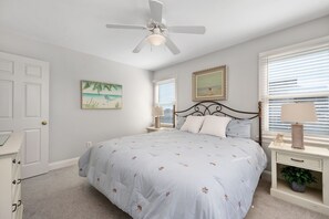 A bedroom with a bed, a ceiling fan, two bedside tables with lamps, a door, and a window with blinds. - Gone Coastal: Private Pool, Hot Tub, Ocean and Sunset Views! (Nags Head)
