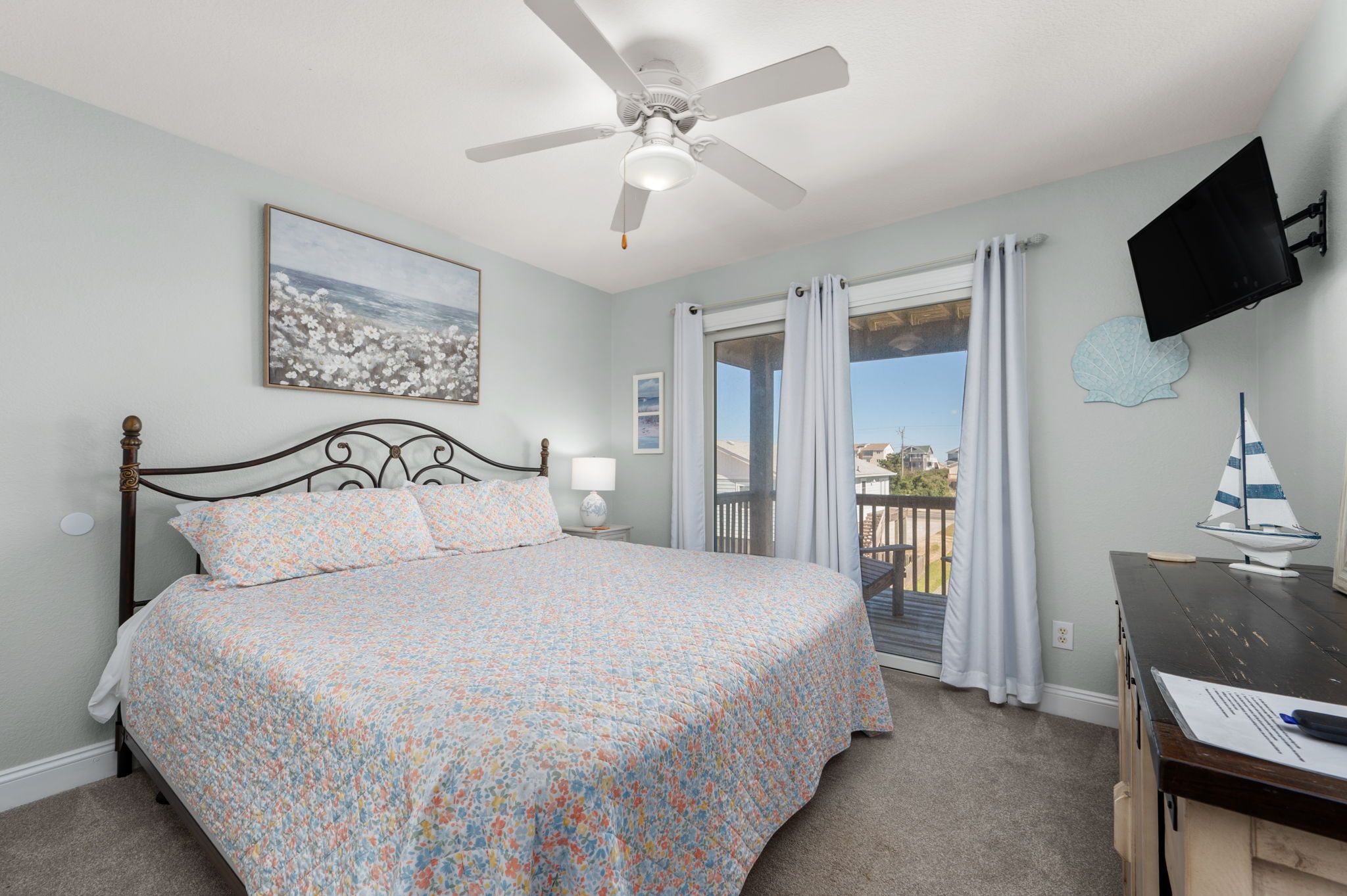 A bedroom with a bed, a ceiling fan, a TV mounted on the wall, a window with curtains, and a view of the outside.