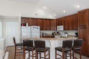 A modern kitchen with wooden cabinets, a central island with bar stools, a refrigerator, microwave, and a coffee maker. - Gone Coastal: Private Pool, Hot Tub, Ocean and Sunset Views! (Nags Head)