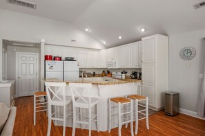 A modern kitchen with white cabinetry, a central island with bar stools, and a stainless steel trash can. - Gone Coastal: Private Pool, Hot Tub, Ocean and Sunset Views! (Nags Head)