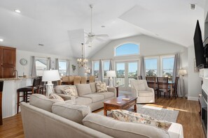 A spacious living area with a high ceiling, a ceiling fan, a fireplace, and large windows offering a view of the outside. - Gone Coastal: Private Pool, Hot Tub, Ocean and Sunset Views! (Nags Head)