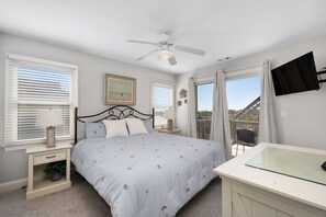 A bedroom with a bed, bedside tables, a ceiling fan, a TV, and a balcony with curtains. - Gone Coastal: Private Pool, Hot Tub, Ocean and Sunset Views! (Nags Head)