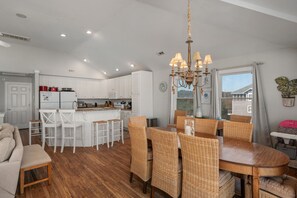 A modern kitchen with white cabinets, a dining area with wicker chairs, and a chandelier. - Gone Coastal: Private Pool, Hot Tub, Ocean and Sunset Views! (Nags Head)