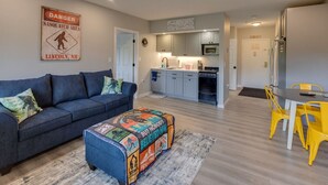 Una sala de estar con un sofá, un pouf colorido y una zona de comedor con sillas amarillas.