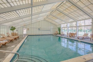Una piscina interior con techos de vidrio transparente, sillas de descanso y plantas en macetas.