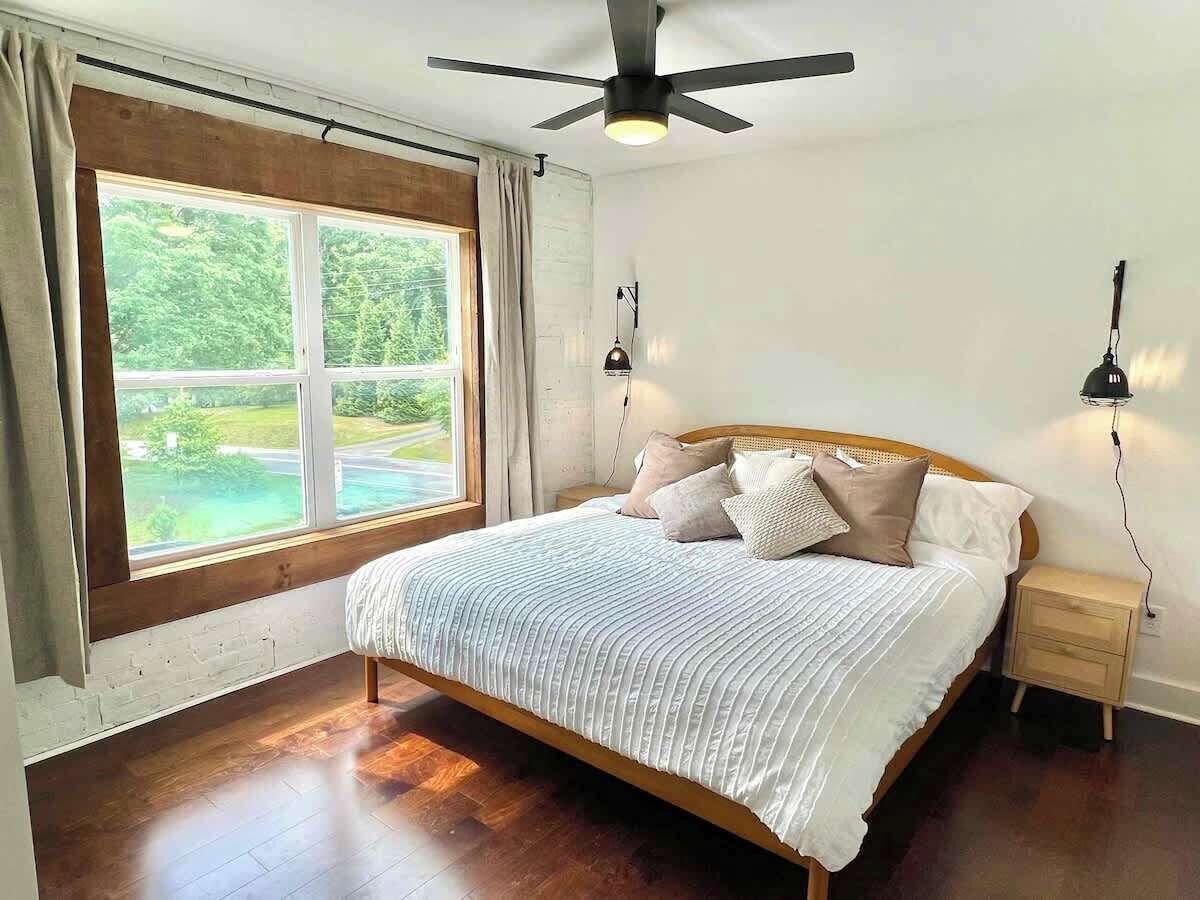 A bedroom with a large window, a bed, a nightstand, a ceiling fan, and wall-mounted lamps.