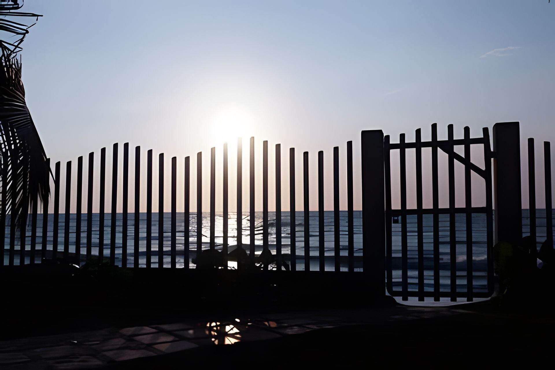 A beach view with a fence in the foreground and the sun setting in the background.